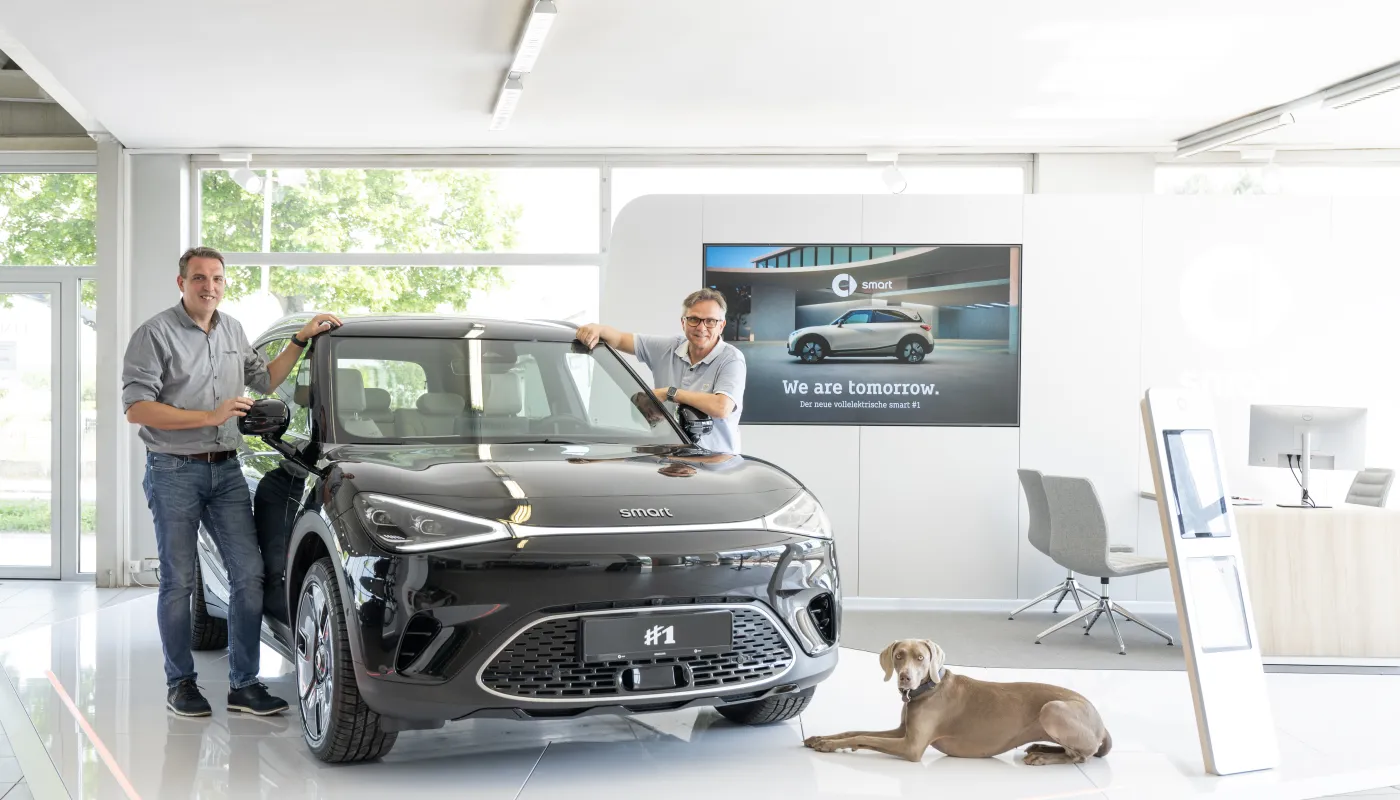 Wolfgang Hummel präsentiert mit einem Verkäufer und Hund ein Auto im Verkaufsraum. 